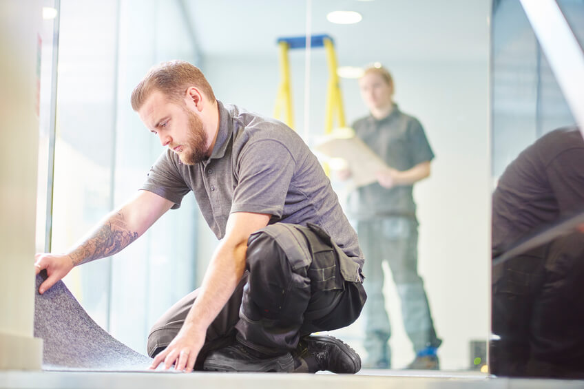 a male carpet fitter is installing carpet in a modern office refurbishment. IN the background a female co-worker can be seen holding plans . They are wearing safety workwear.